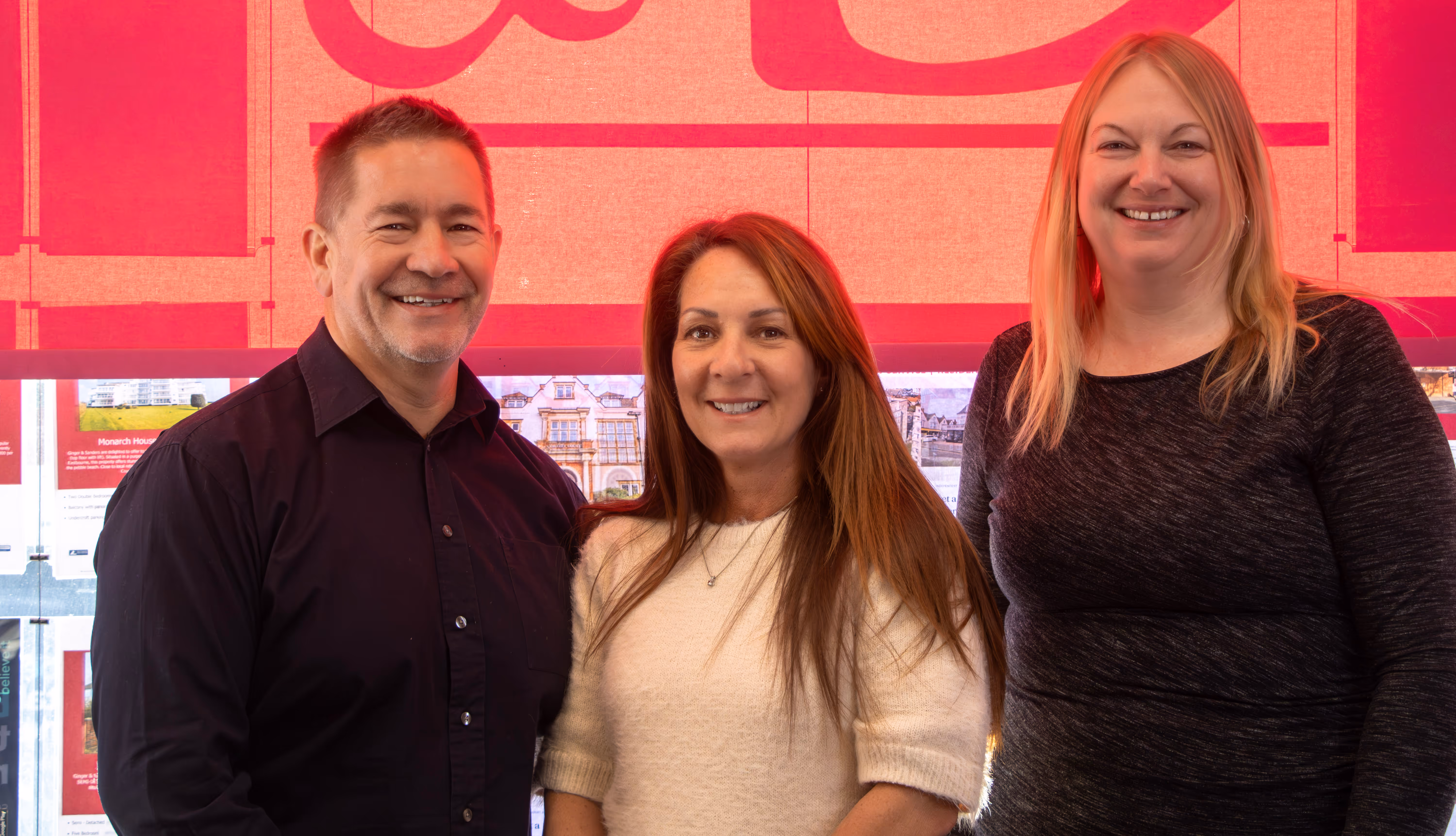 Three people smiling while standing together indoors against a pink and red background.