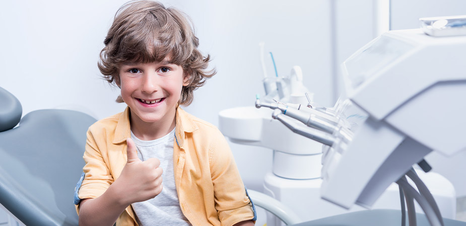 A young child sitting in a dentist's chair, giving a thumbs up, surrounded by dental equipment in a bright, modern office.