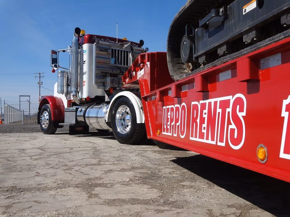 Close-up of a red and white heavy-duty truck with a red trailer carrying construction equipment labeled 'LEPPO RENTS'.