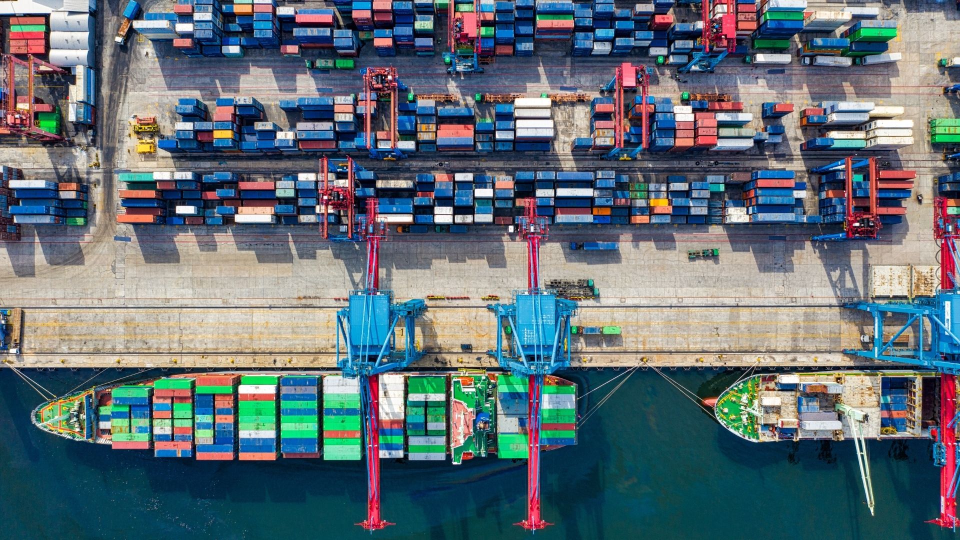 Aerial view of a busy shipping port with colorful stacked containers and cranes. Two cargo ships are docked, indicating active industrial activity.