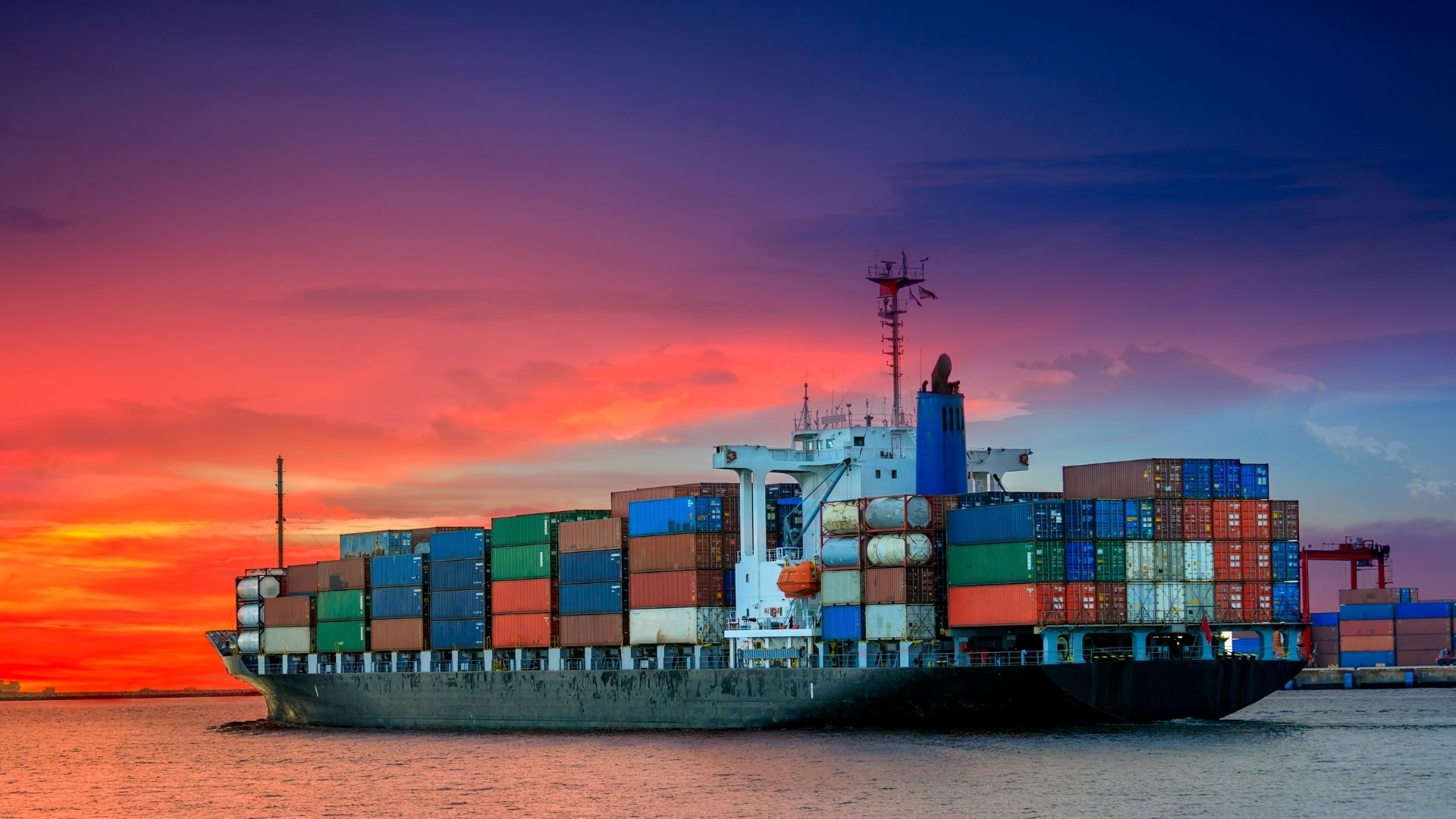 A large cargo ship loaded with colorful containers sails on calm waters at sunset. The sky is vibrant with orange, pink, and blue hues, creating a serene atmosphere.
