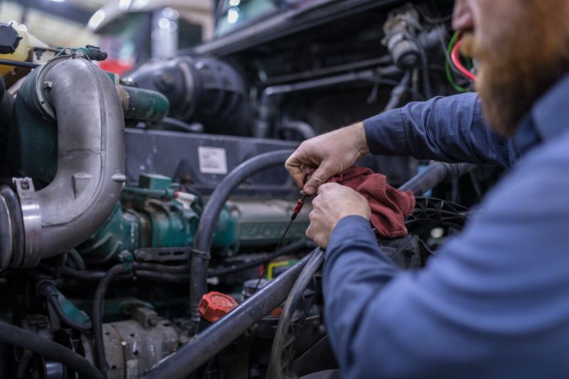 Mechanic inspecting diesel truck engine, wiping oil dipstick with red rag, surrounded by hoses, turbocharger, and various engine components in shop. 