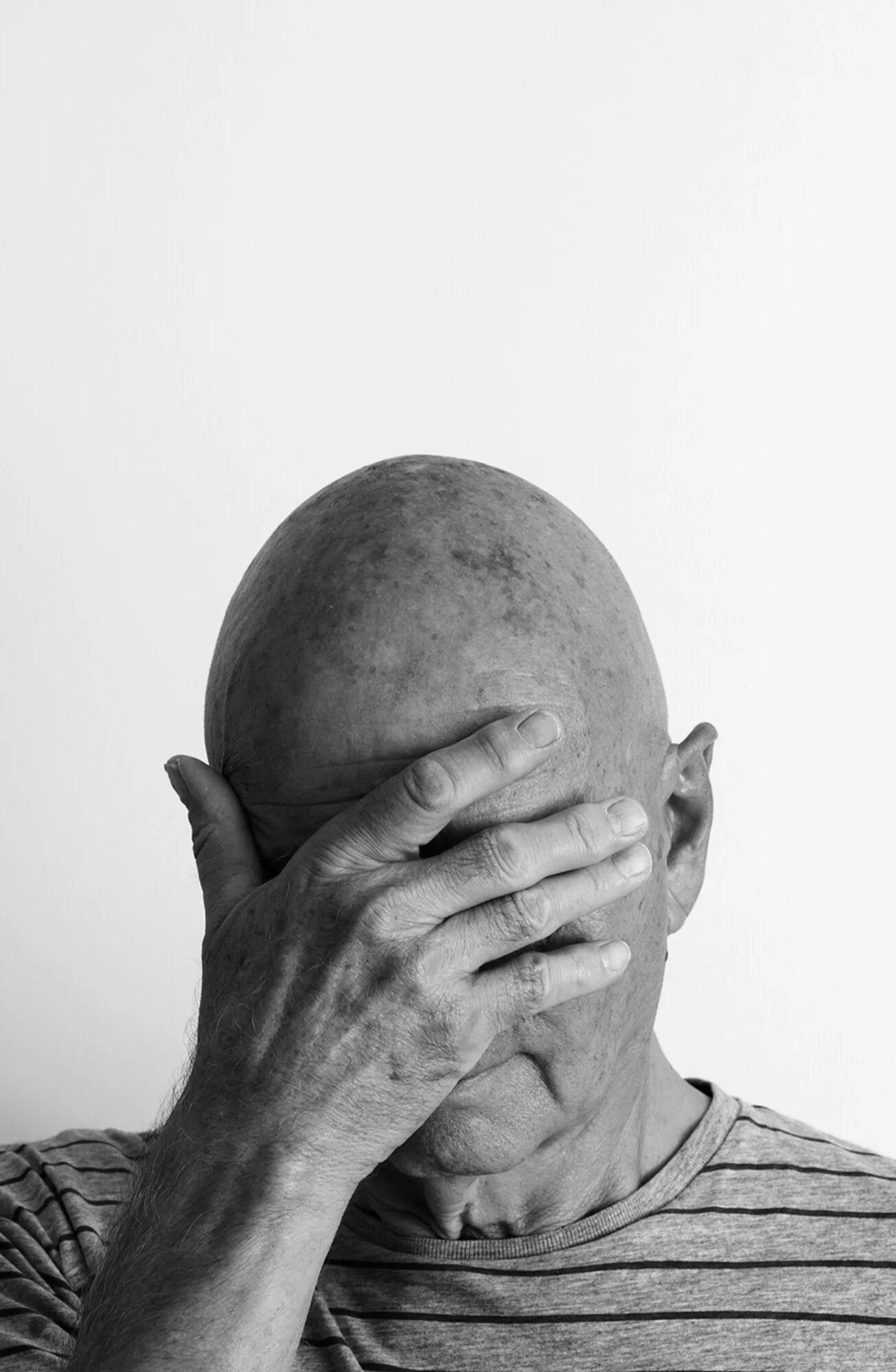 Marcel Peltier – Werkserie „Darkness“ (2022): Schwarz-Weiß-Porträt eines kahlköpfigen Mannes mit gestreiftem Hemd, der seine Hand vor sein Gesicht hält.