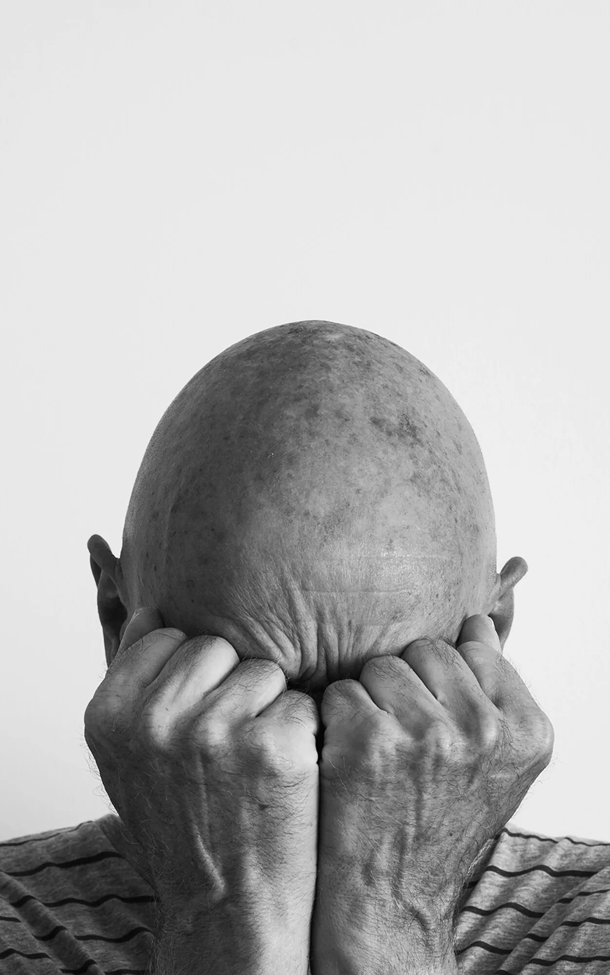 Schwarz-Weiß-Foto eines kahlköpfigen Mannes, der sein Gesicht in seinen geballten Fäusten vergräbt und damit Verzweiflung oder Frustration ausdrückt.