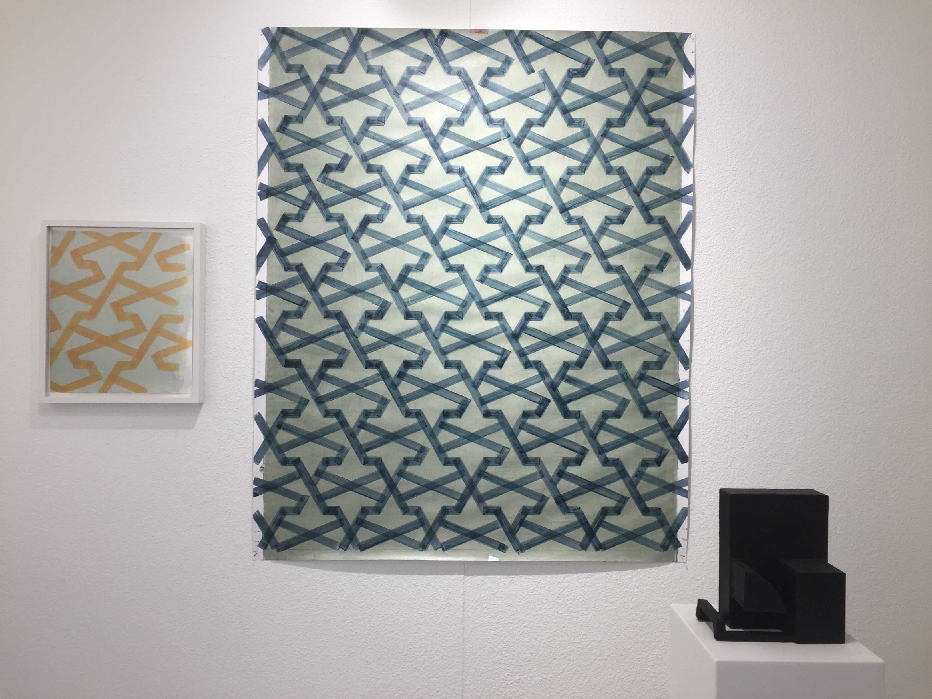 Ausstellungsansicht im Kunstverein Olten 2017: Zwei abstrakte ornamentale Bilder von Marcel Peltier hängen an einer weißen Wand, links ein kleineres orangefarbenes Motiv, daneben ein größeres blau-grünes Musterbild. Rechts davor steht ein schwarzes Objekt von Markus Wyss auf einem Sockel.
