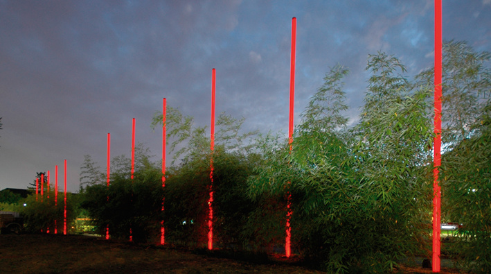 Installation struktur 1 am Kantonsspital Olten: Rote LED-Stelen in gleichmäßiger Reihe entlang eines beleuchteten Weges neben einer Bambushecke, aufgenommen in der Dämmerung mit Gebäuden im Hintergrund.