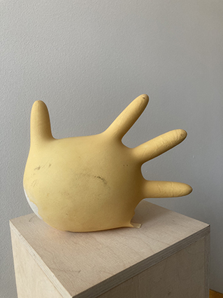 Gelbe, organisch anmutende Polyesterform mit mehreren fingerartigen Ausstülpungen, liegend auf einem Holzsockel präsentiert.