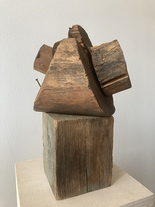 Abstrakte Skulptur aus verschiedenen Holzstücken, asymmetrisch zu einem blockartigen Körper zusammengestellt und auf einem Holzsockel platziert.