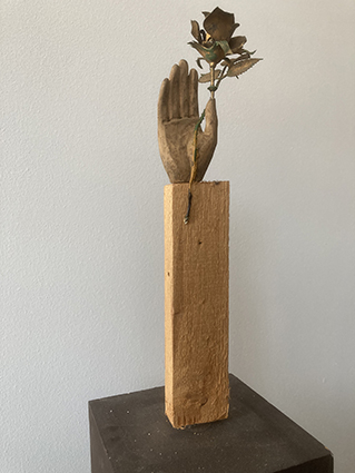 Schmale Holzskulptur mit einer aufrecht stehenden geschnitzten Hand, die oben einen kleinen Metallzweig mit Blättern hält.