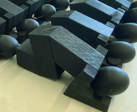 Marcel Peltier – "Praying" Modell eines unrealisierten Projekts (2023): Dreidimensionale Modellarbeit aus dunklem Holz oder holzähnlichem Material auf hellem Untergrund. Mehrere abstrahierte, blockhafte Figuren mit kugelförmigen Köpfen sind in dichter Anordnung dargestellt. Die Körper bestehen aus kantigen, geometrischen Volumen, die in gebeugter, nach vorne geneigter Haltung angeordnet sind und eine kollektive, beinahe rituelle Bewegung andeuten. Die reduzierte Formensprache und das serielle Nebeneinander der Figuren verleihen dem Modell eine stille, kontemplative und zugleich architektonisch strukturierte Wirkung.