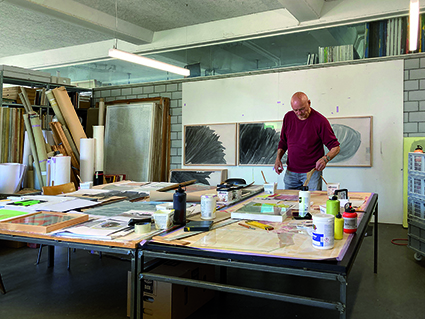 Marcel Peltier in seinem Atelier in Olten. Vordergrund ist ein Tisch mit Farben, Leinwänden etc. zu sehen und im Hintergrund hängen Bilder der Serie "Les fleurs du mal" an einer weissen Wand. 