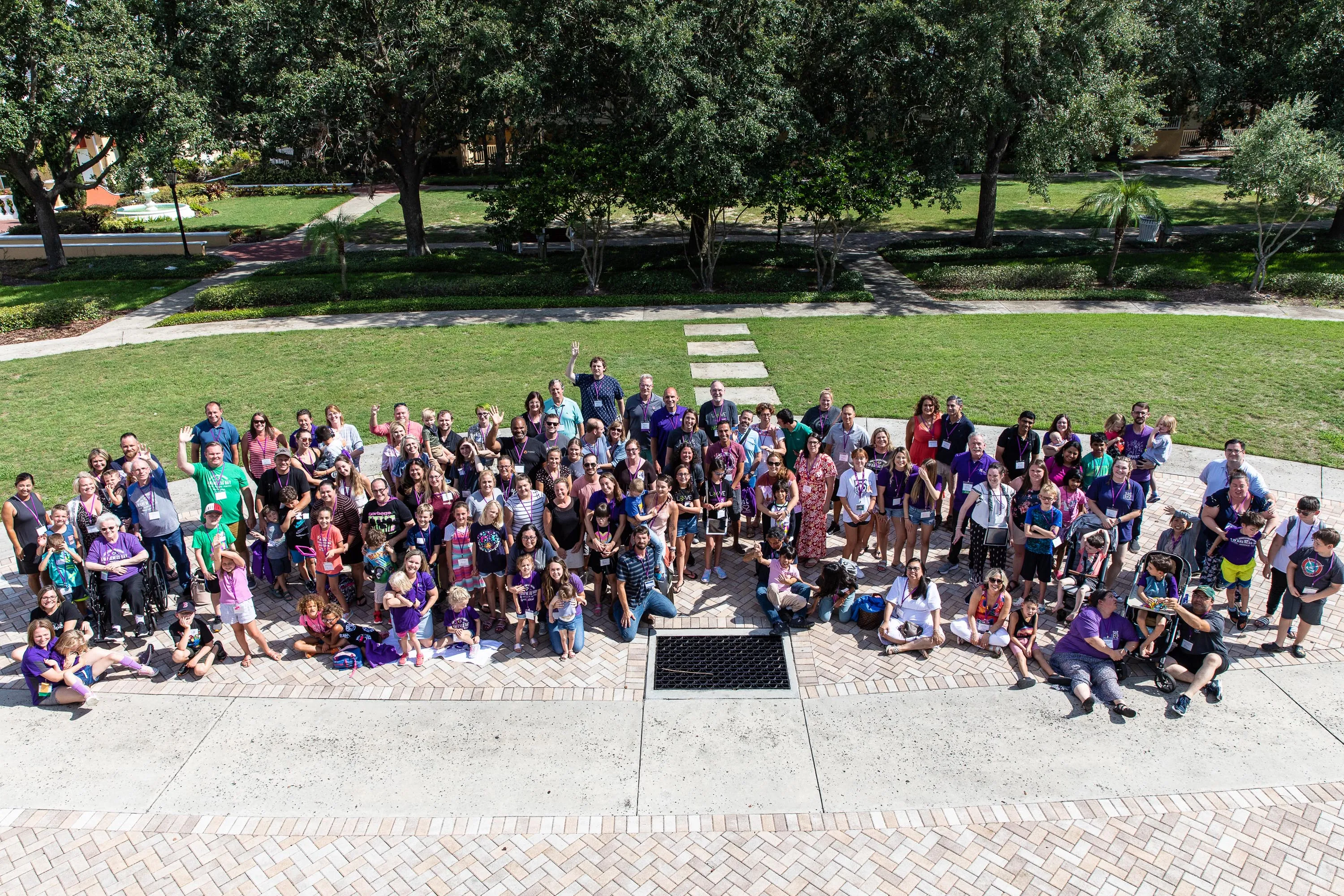 Large diverse group of people gathered outdoors on a paved area with green grass and trees in the background, many smiling and waving at PEC Field Day.
