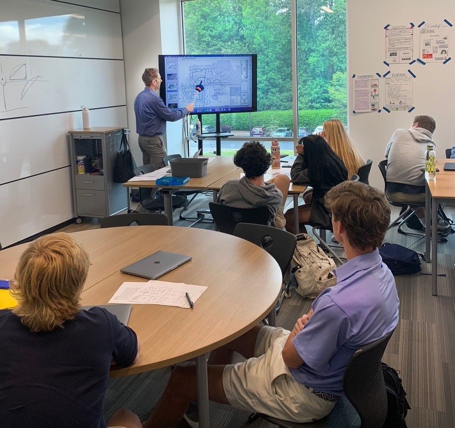 Ken, PEC CEO, teaching about architecture and engineering, pointing at a large screen with a map diagram while students sit at tables in a classroom with large windows showing green trees outside.
