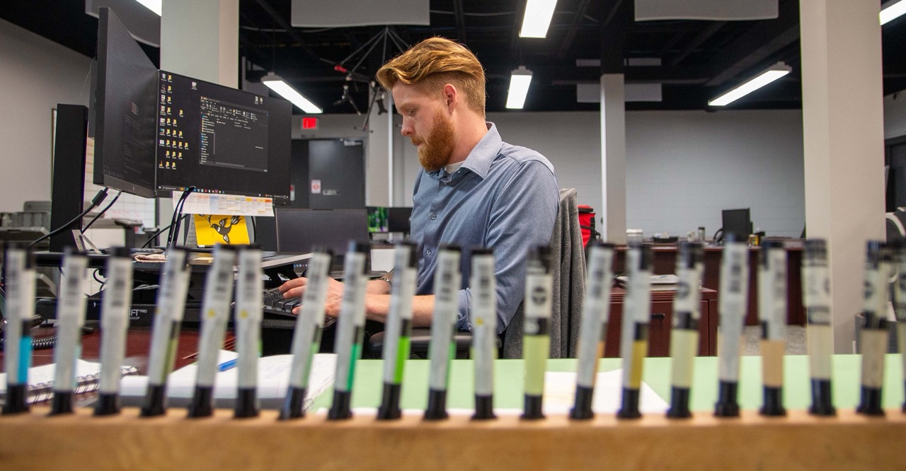 PEC employee working on a computer at an office desk, viewed behind a row of blurred landscape architecture pens.