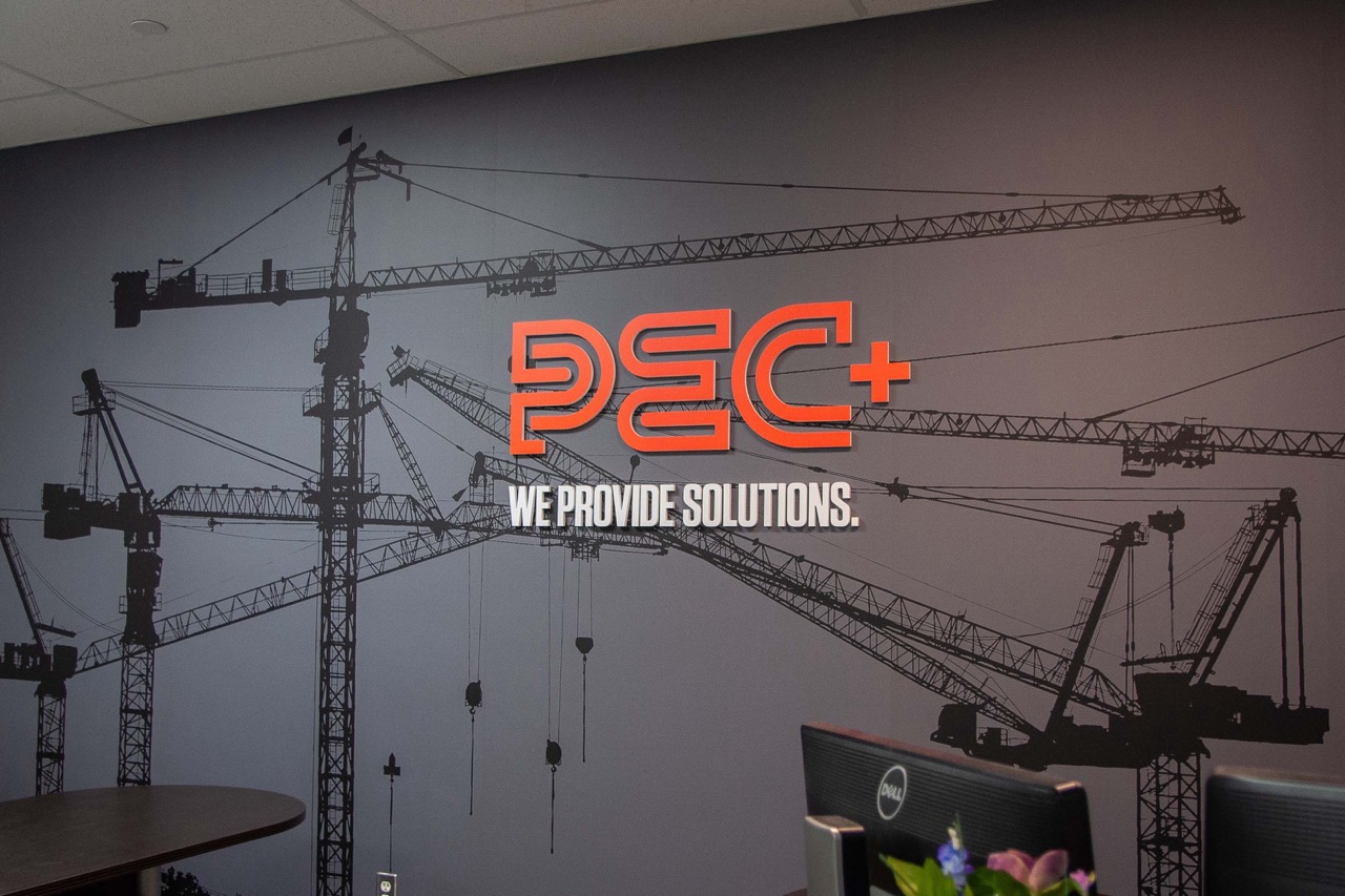 Office wall with black crane silhouettes and bright orange PEC+ logo with the text 'We Provide Solutions.'