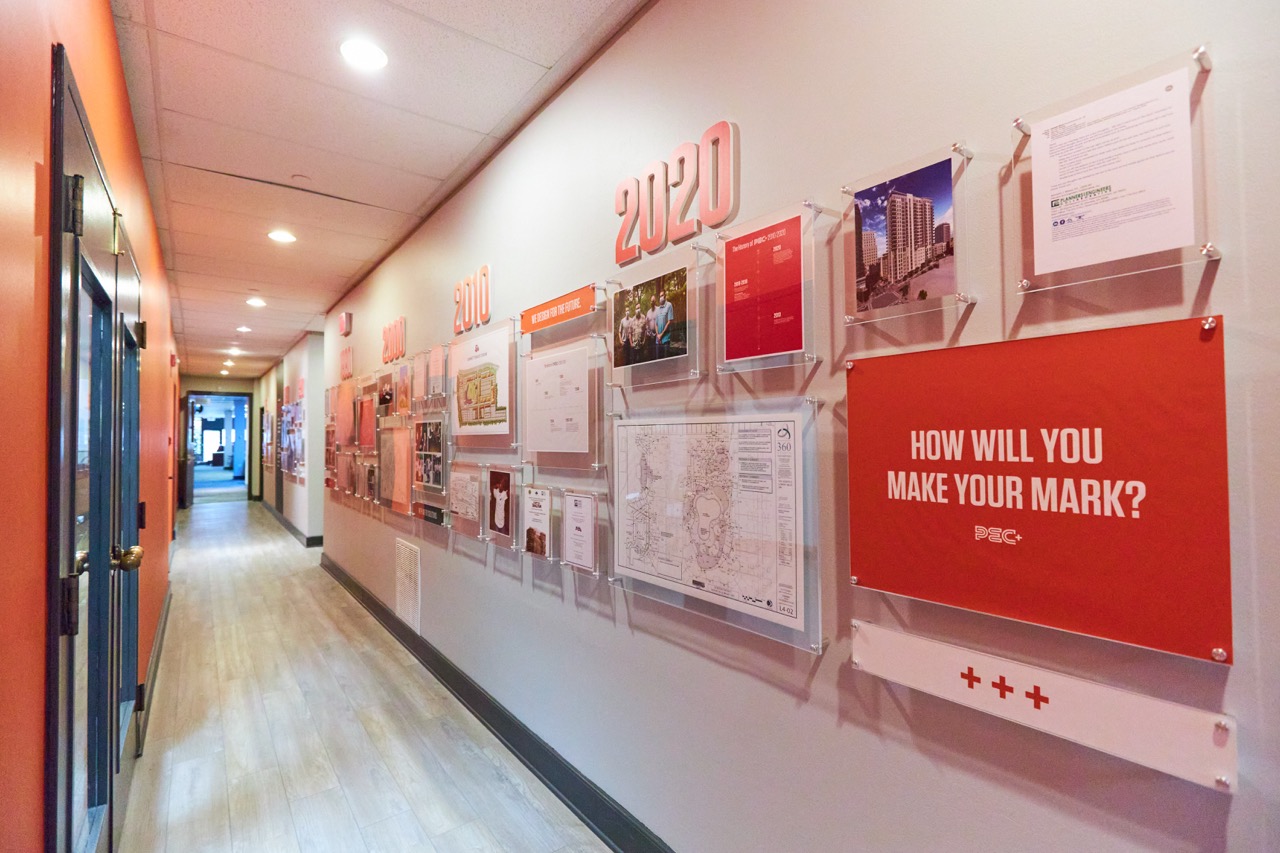 PEC Office hallway with a timeline display on the wall showcasing the 60+ year history of PEC, including the years 2000, 2010, and 2020. The display features photos, maps, and a sign that reads "How will you make your mark?"
