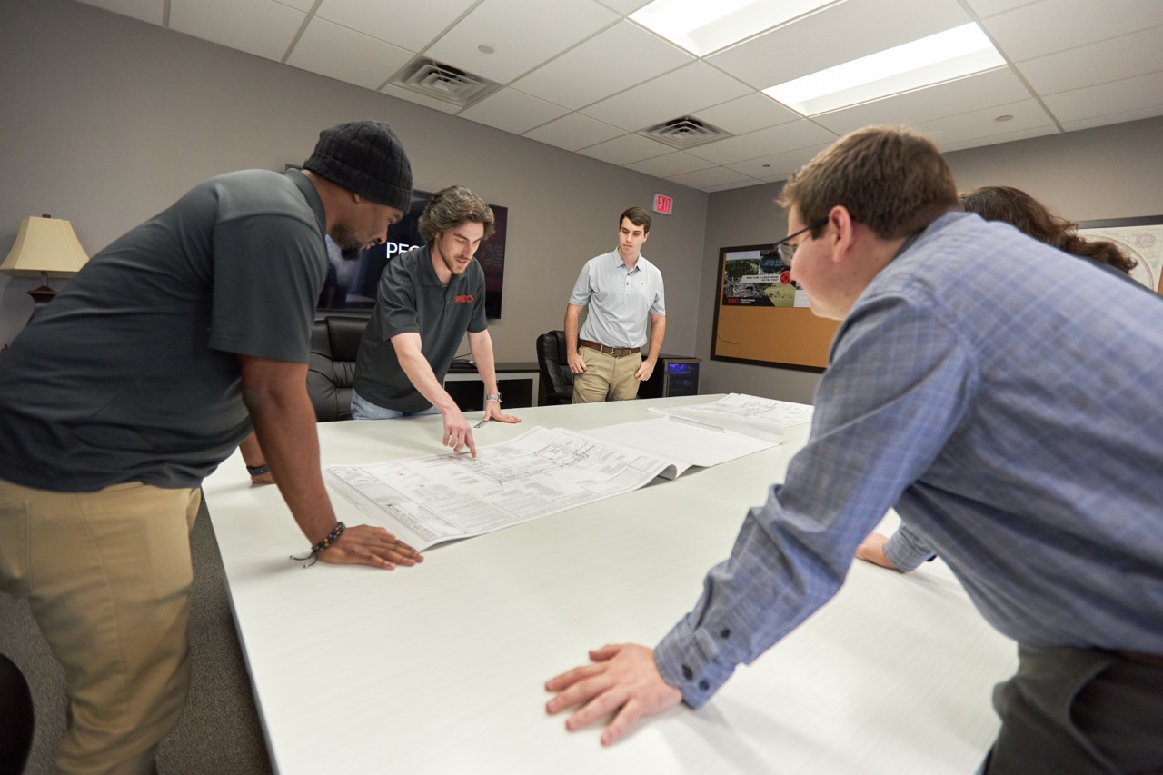 Five PEC employees gathered around a table reviewing large architectural blueprints in the PEC conference room. 