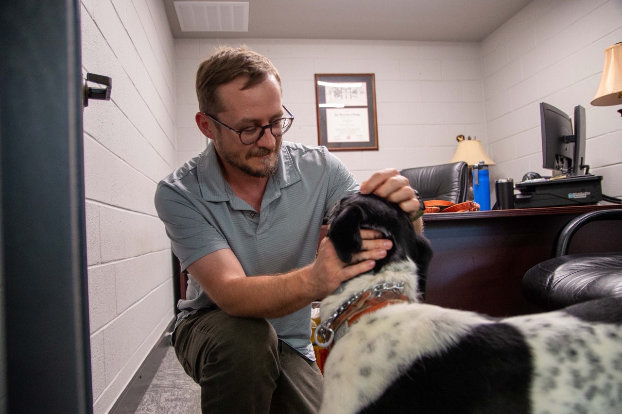 Man wearing glasses and a gray polo shirt petting a black and white dog inside an office showing PEC offices are a pet-friendly.