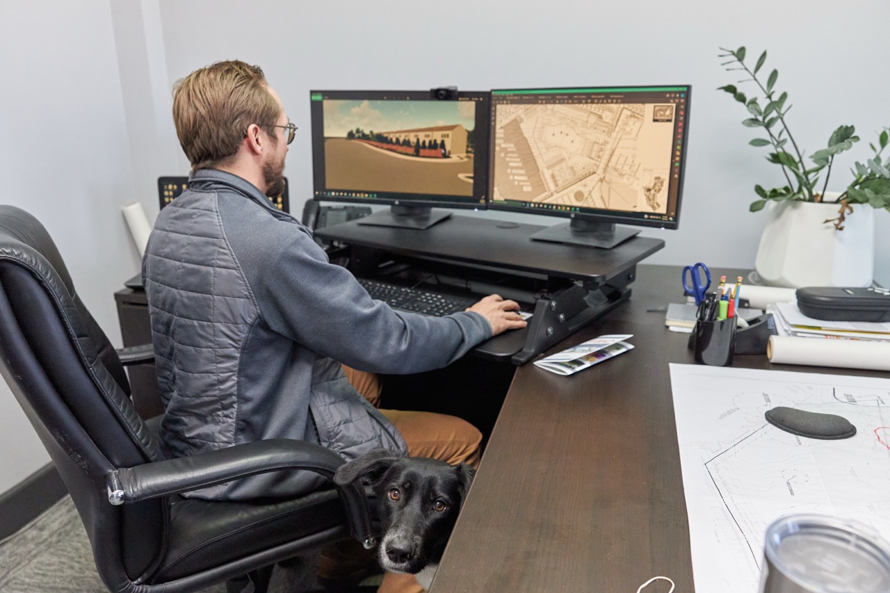 Man working on an engineering sketch at a desk with a black dog peeking from underneath the desk.