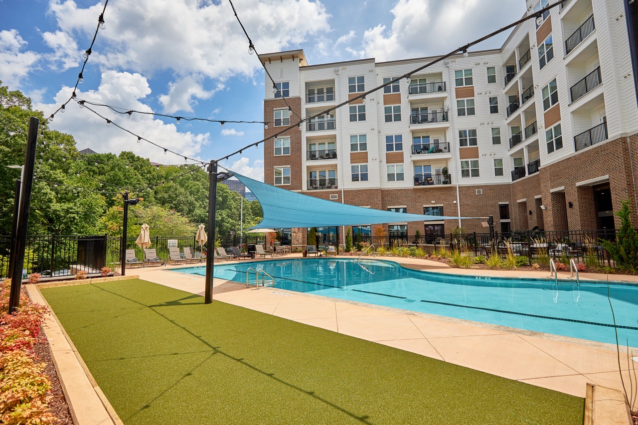 Outdoor pool area with sunshade, apartment building, trees, and a partly cloudy sky, highlighting PEC+ services, Landscape Architecture and Land Planning.
