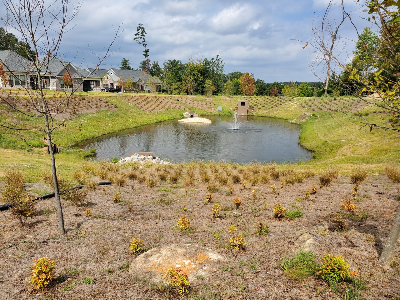 Man-made pond with a water fountain surrounded by grassy banks and small shrubs, with houses and trees in the background under a partly cloudy sky PEC service Water Resources.