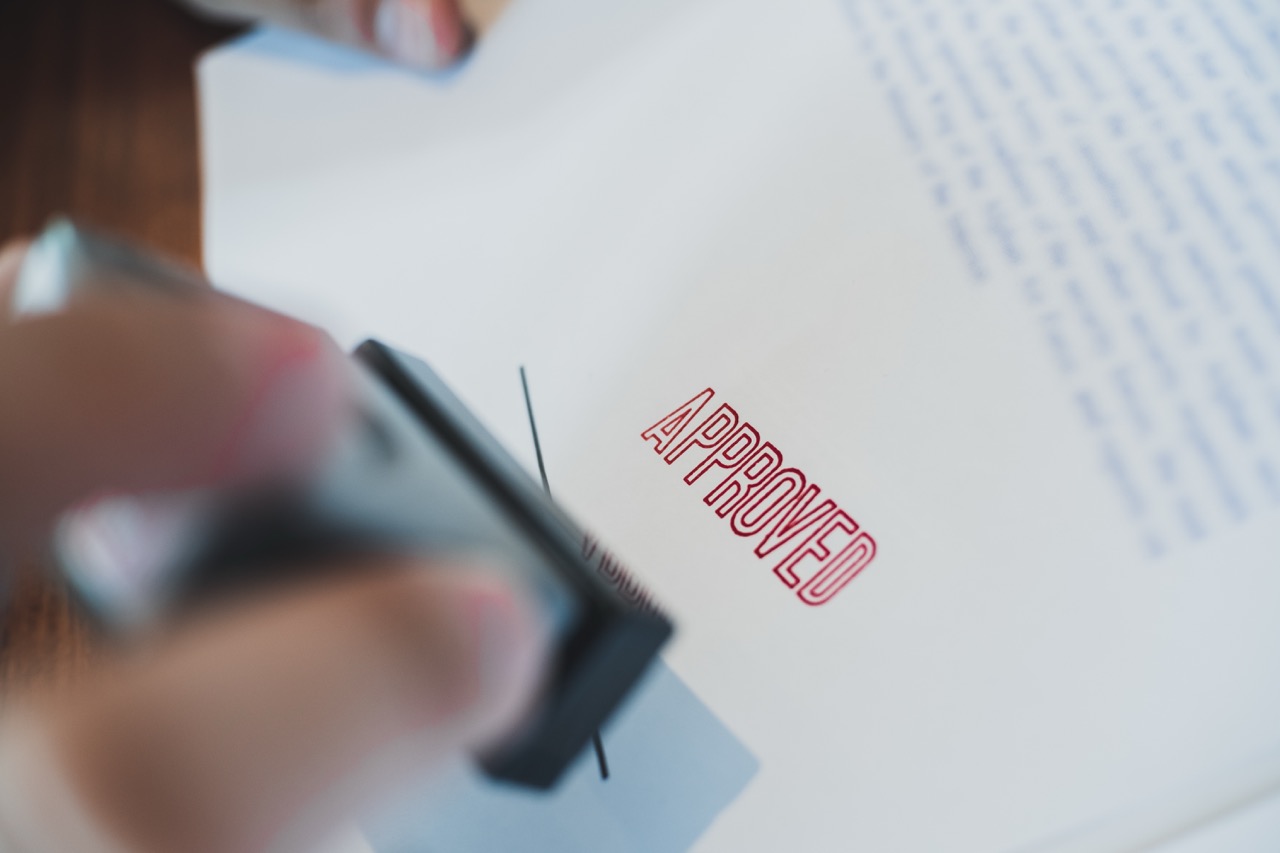 Close-up of a hand stamping a document with a red "APPROVED" stamp showing PEC service Permit Expediting.