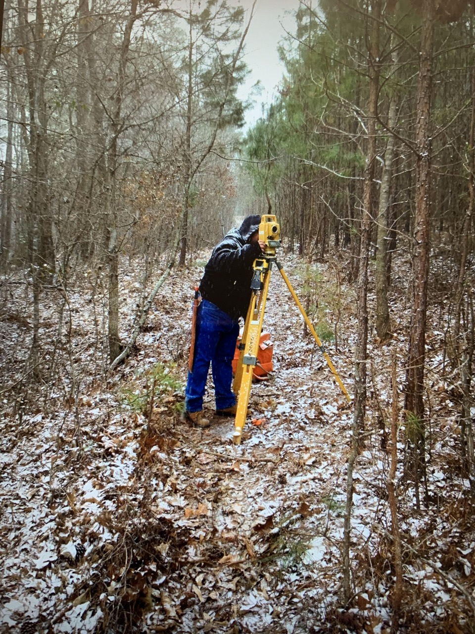 PEC employee in winter clothing operating a yellow surveying instrument on a tripod in a forest with light snow on the ground showing PEC service Surveying.