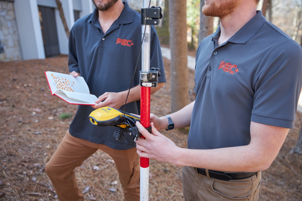 Two PEC employees wearing gray PEC+ polo shirts outdoors with surveying equipment and a notebook showing PEC service Surveying.