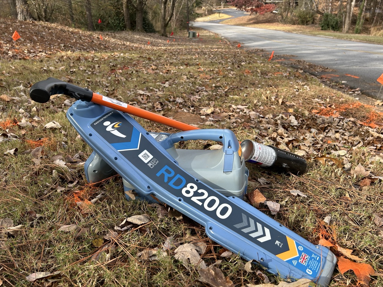 RD8200 utility locating device resting on grass and fallen leaves beside a roadside marked with orange flags and paint showing PEC Utility Locate Field service.