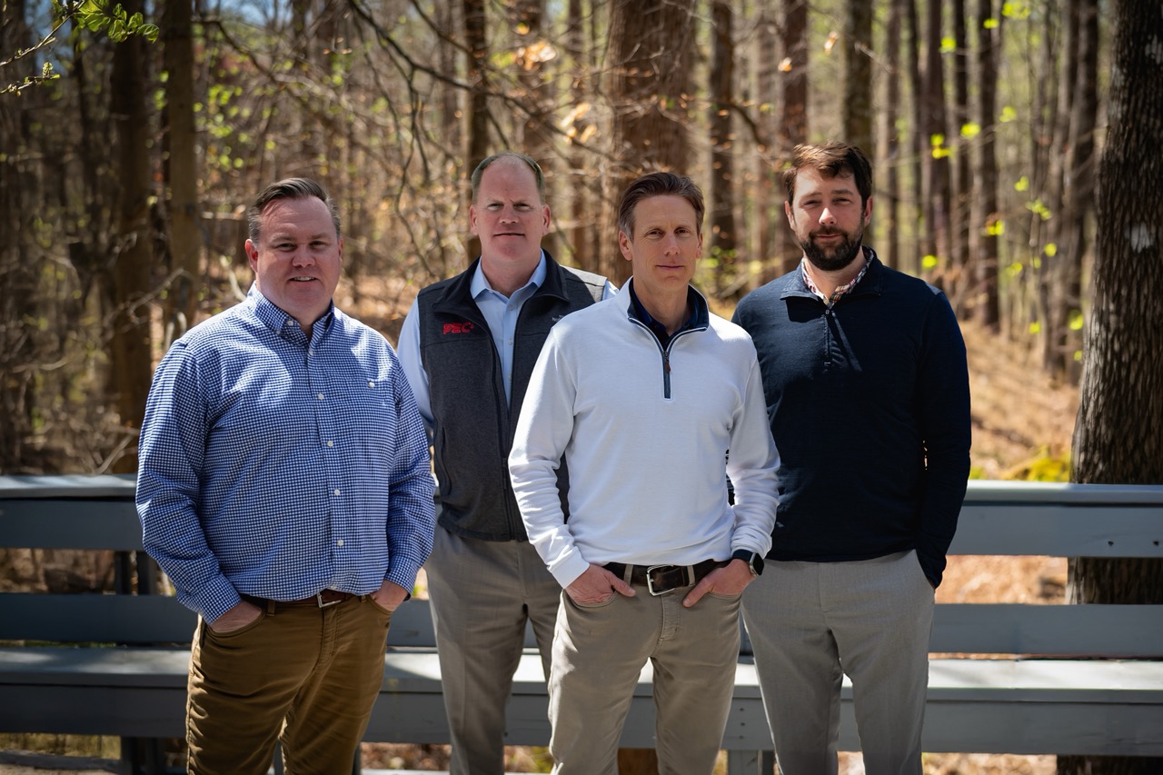 Four men comprising PEC Leadership standing outdoors in front of a wooden fence with trees in the background, dressed in casual business attire.