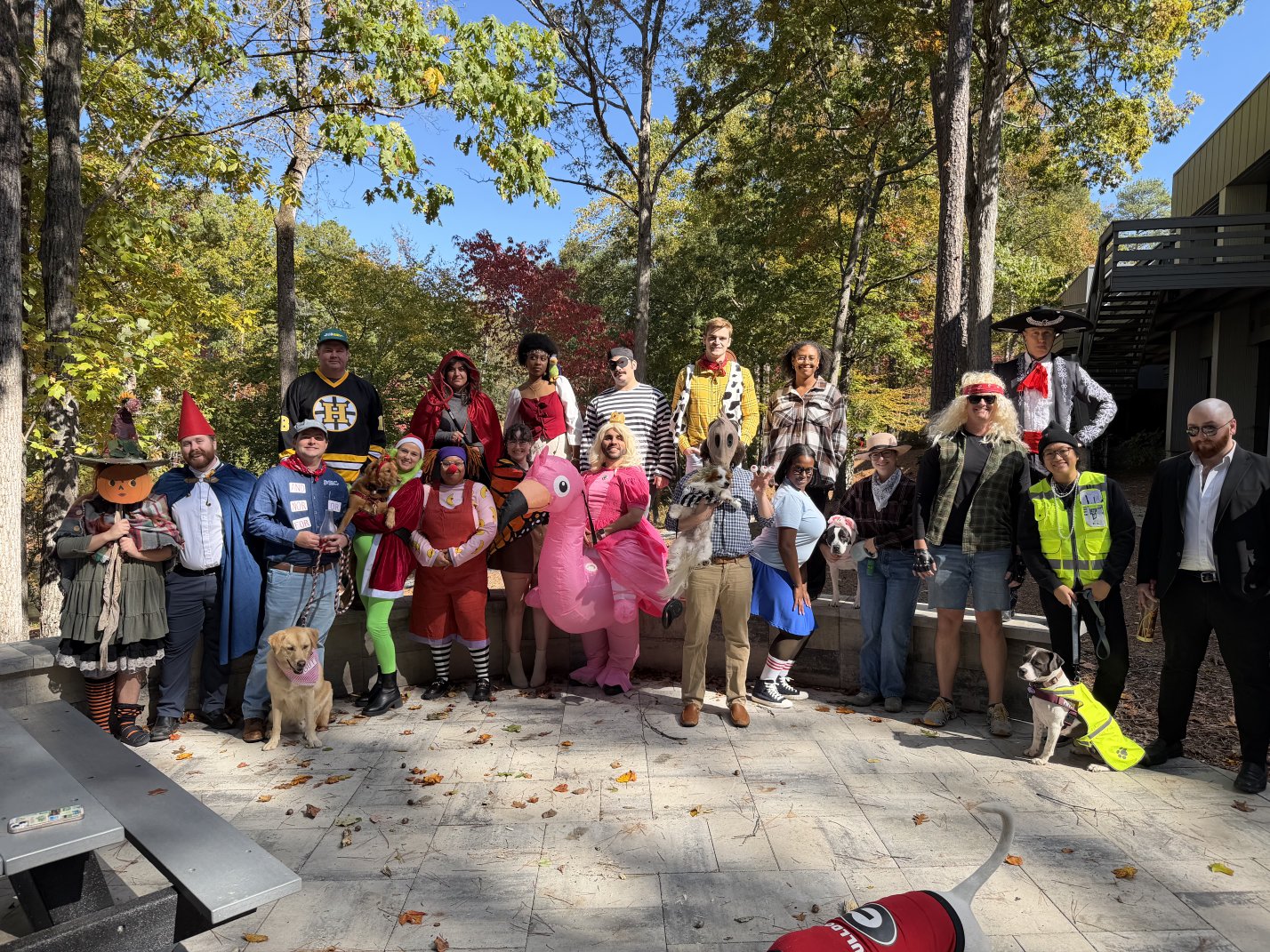 Group of PEC employees outdoors dressed in various colorful costumes, some holding dogs, standing on a stone pavement with trees in the background for PEC Halloween Costume Contest.