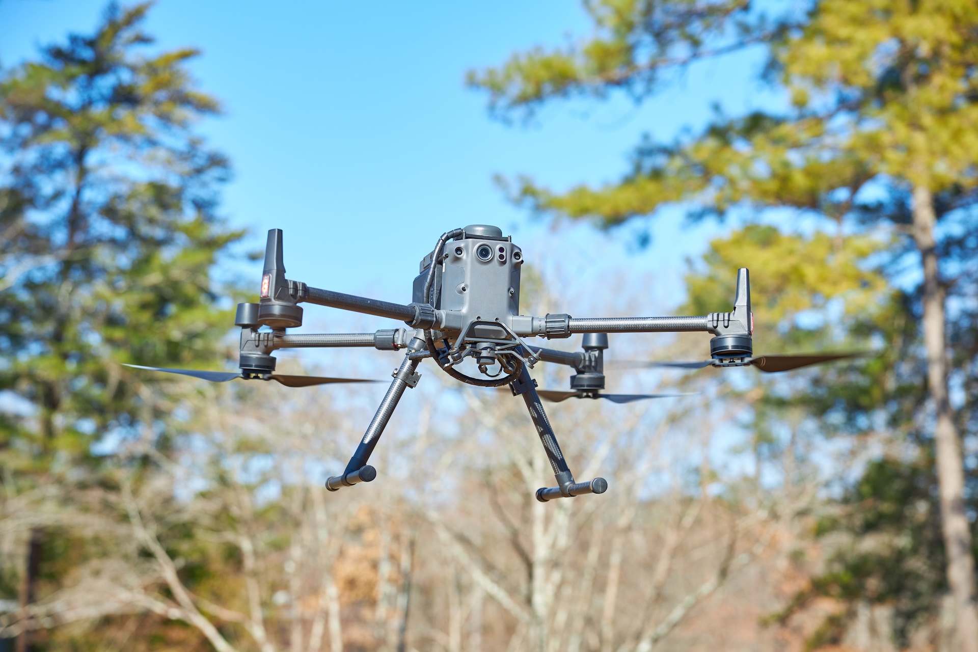 Close-up of a quadcopter drone with carbon fiber arms flying outdoors against a background of trees and blue sky showing PEC Aerial Field service.