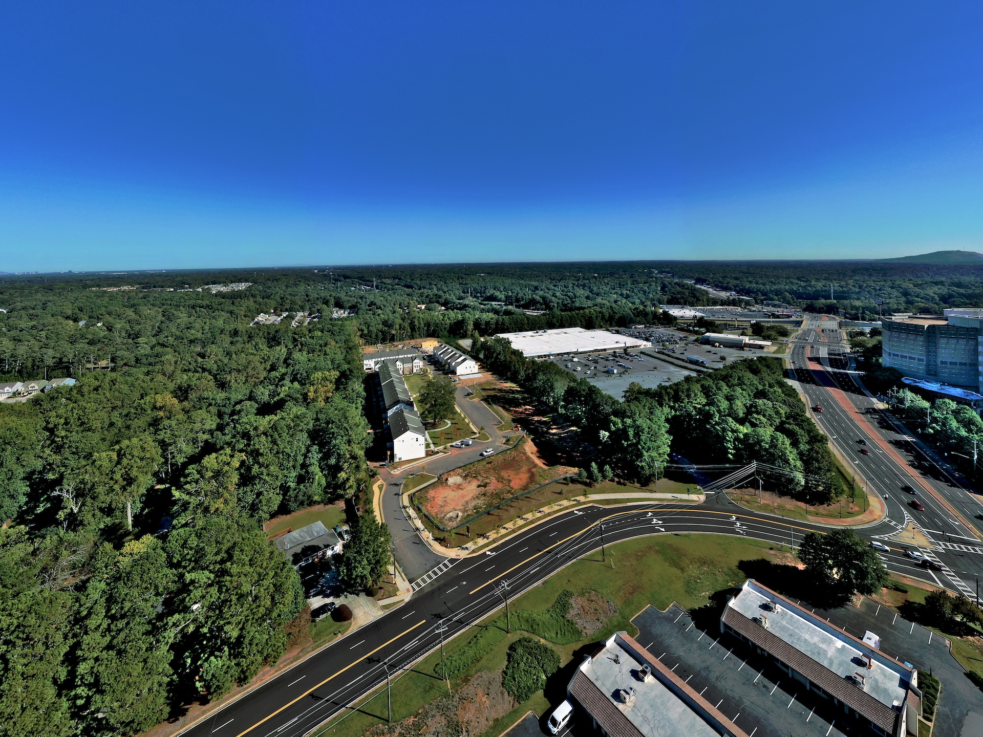 Aerial view of a suburban area with dense green trees, residential buildings, parking lots, and a multi-lane road under a clear blue sky showing PEC Aerial Field service.