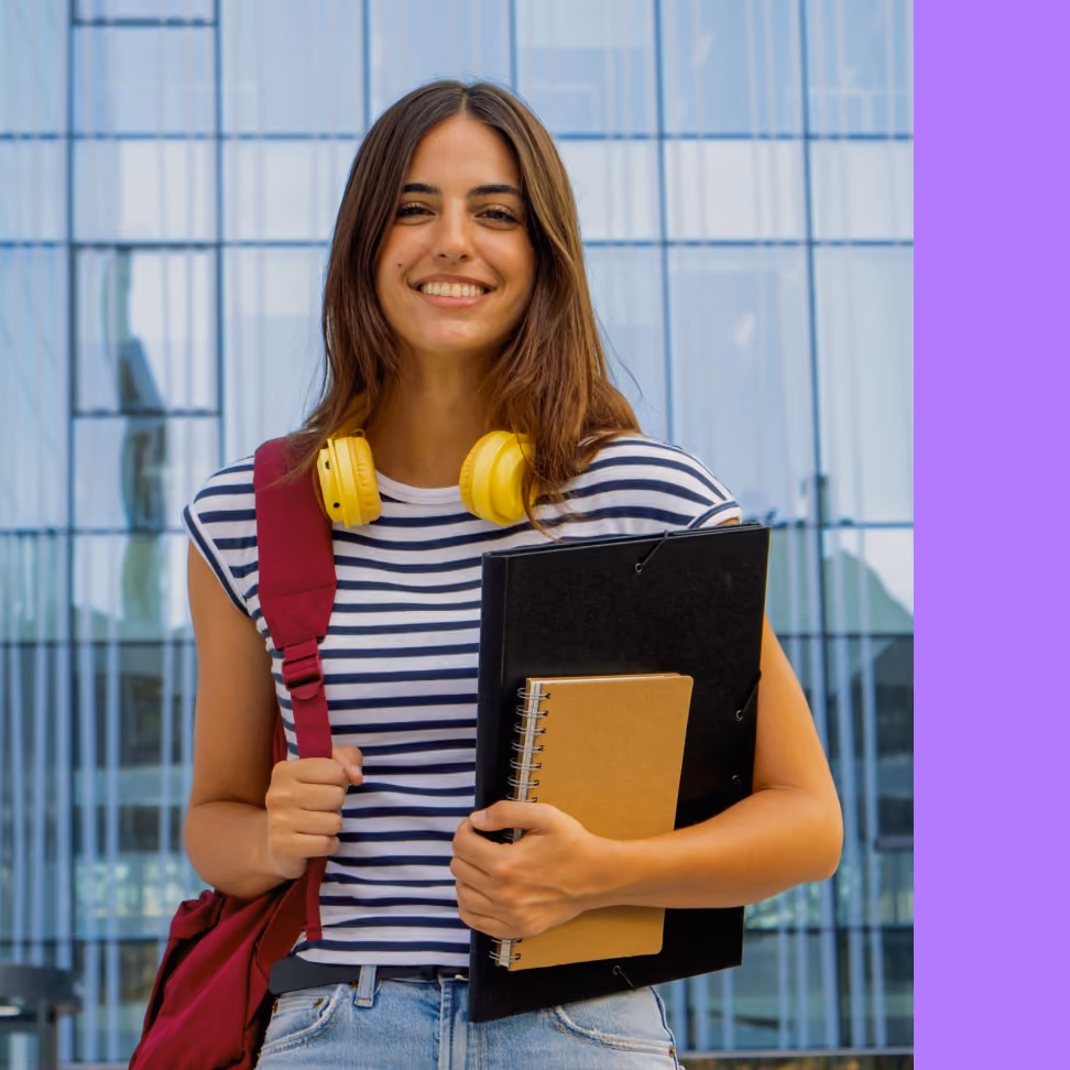 A young woman with long brown hair, wearing a striped shirt, yellow headphones around her neck, and a red backpack, is standing outside a modern glass building. She is holding a black folder and a spiral notebook, smiling confidently at the camera.