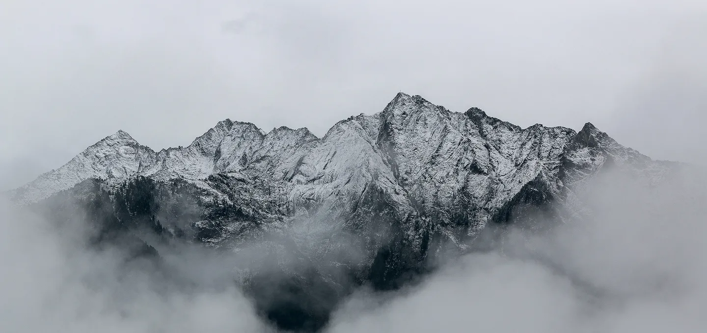 Snow-covered mountain peaks partially obscured by low-hanging fog under a gray sky.