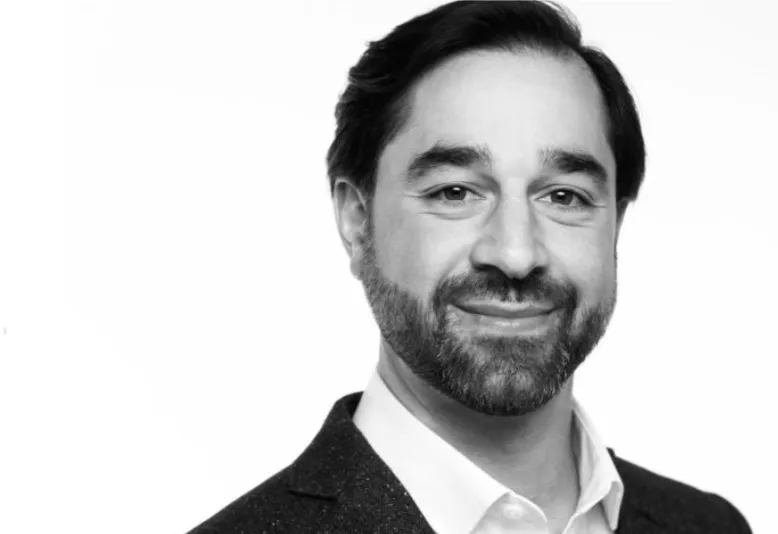 Black and white headshot of a smiling man with dark hair, beard, wearing a suit jacket and white shirt.