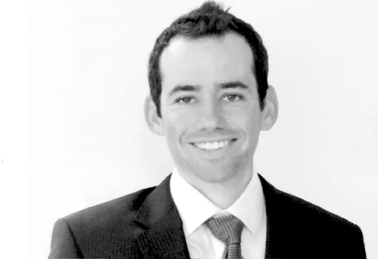 Man in a dark suit, white shirt, and tie smiling with a plain light background.