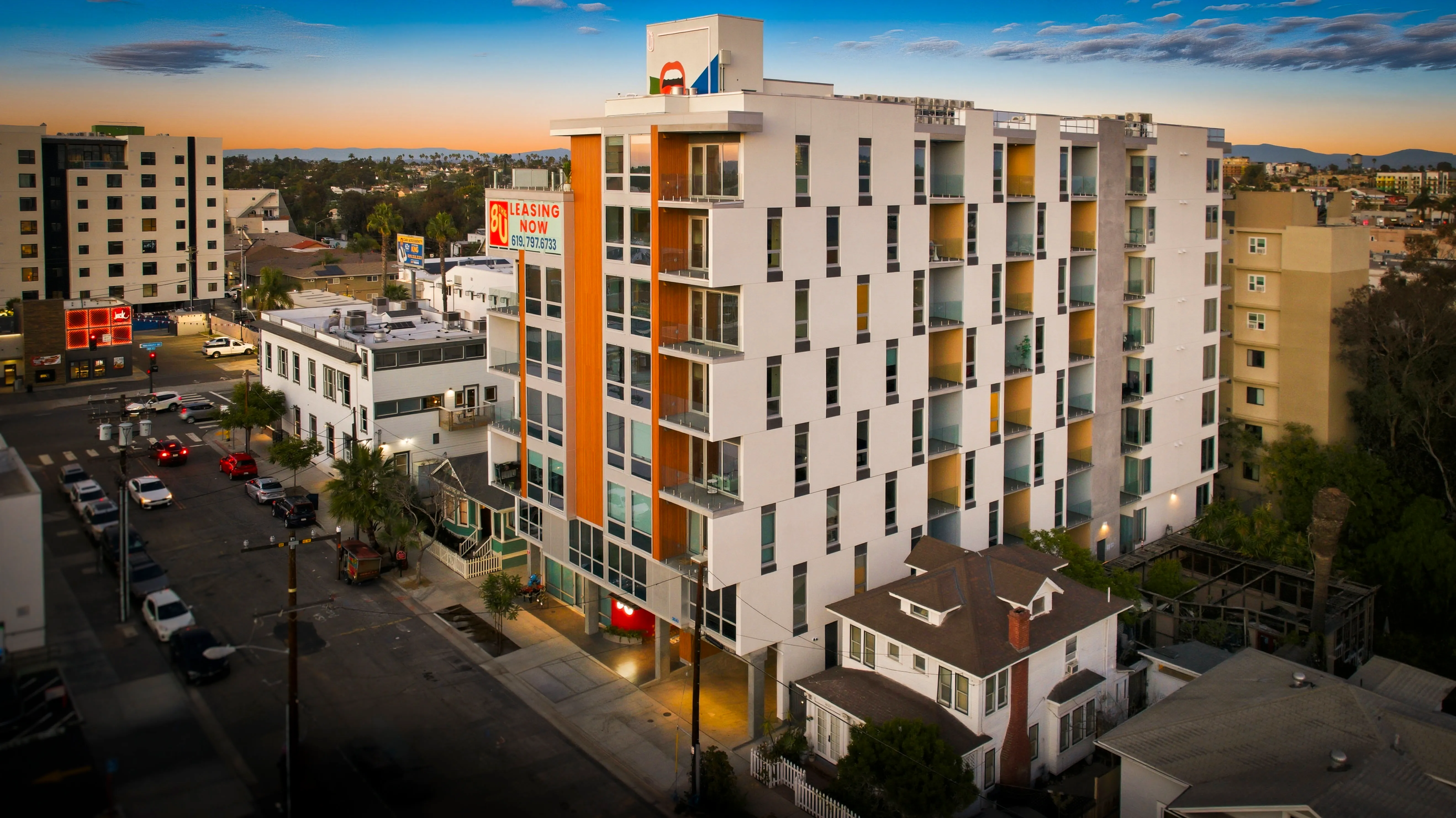 Modern multi-story apartment building with balconies and a leasing sign at sunset in an urban area.