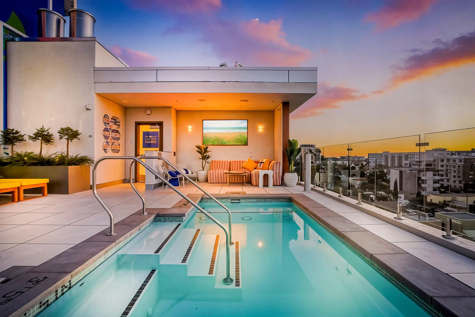 Rooftop infinity pool with seating area and glass railing overlooking cityscape at sunset.