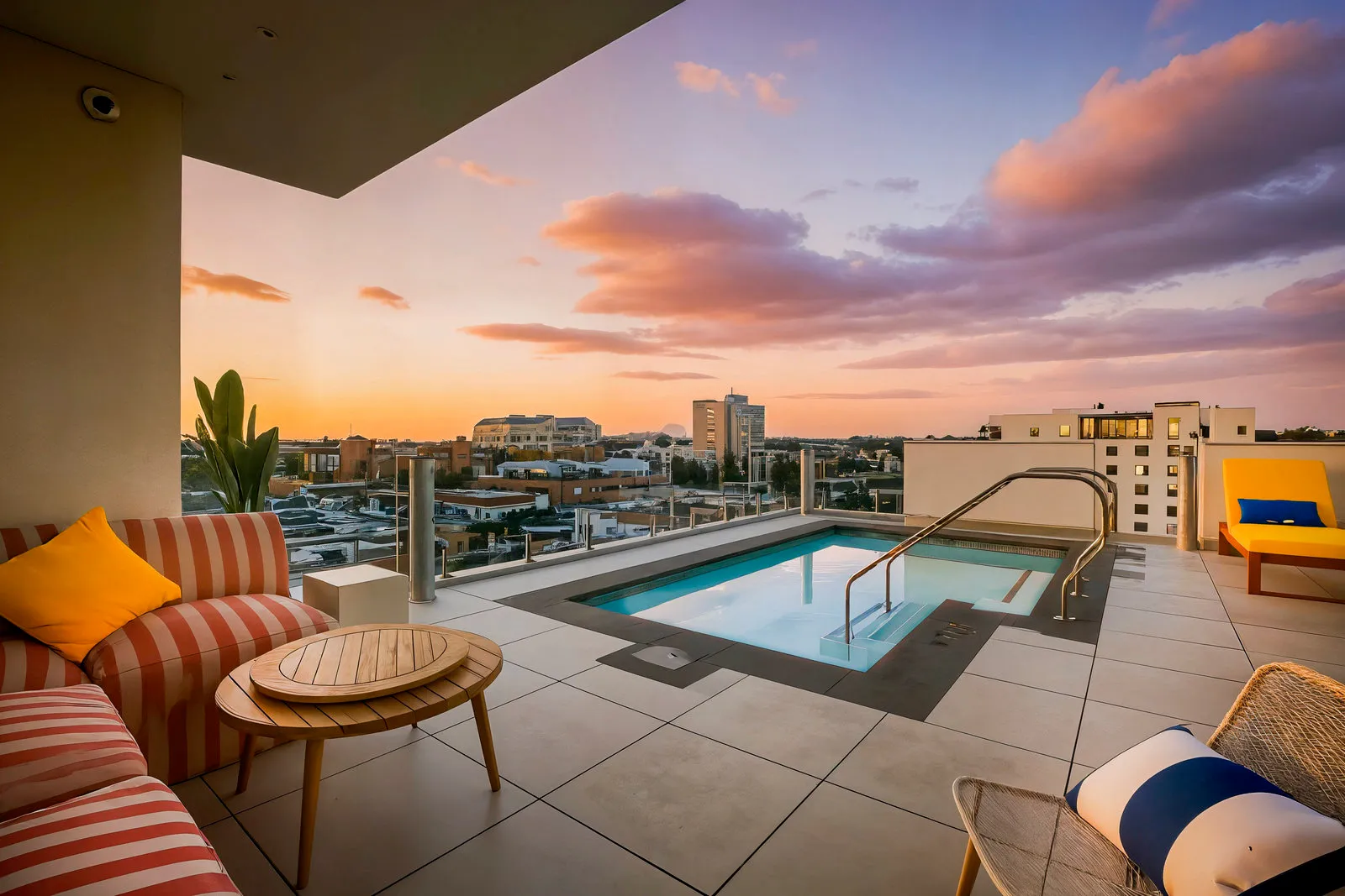 Modern rooftop patio with pool, striped sofa, yellow lounge chair, and cityscape at sunset.