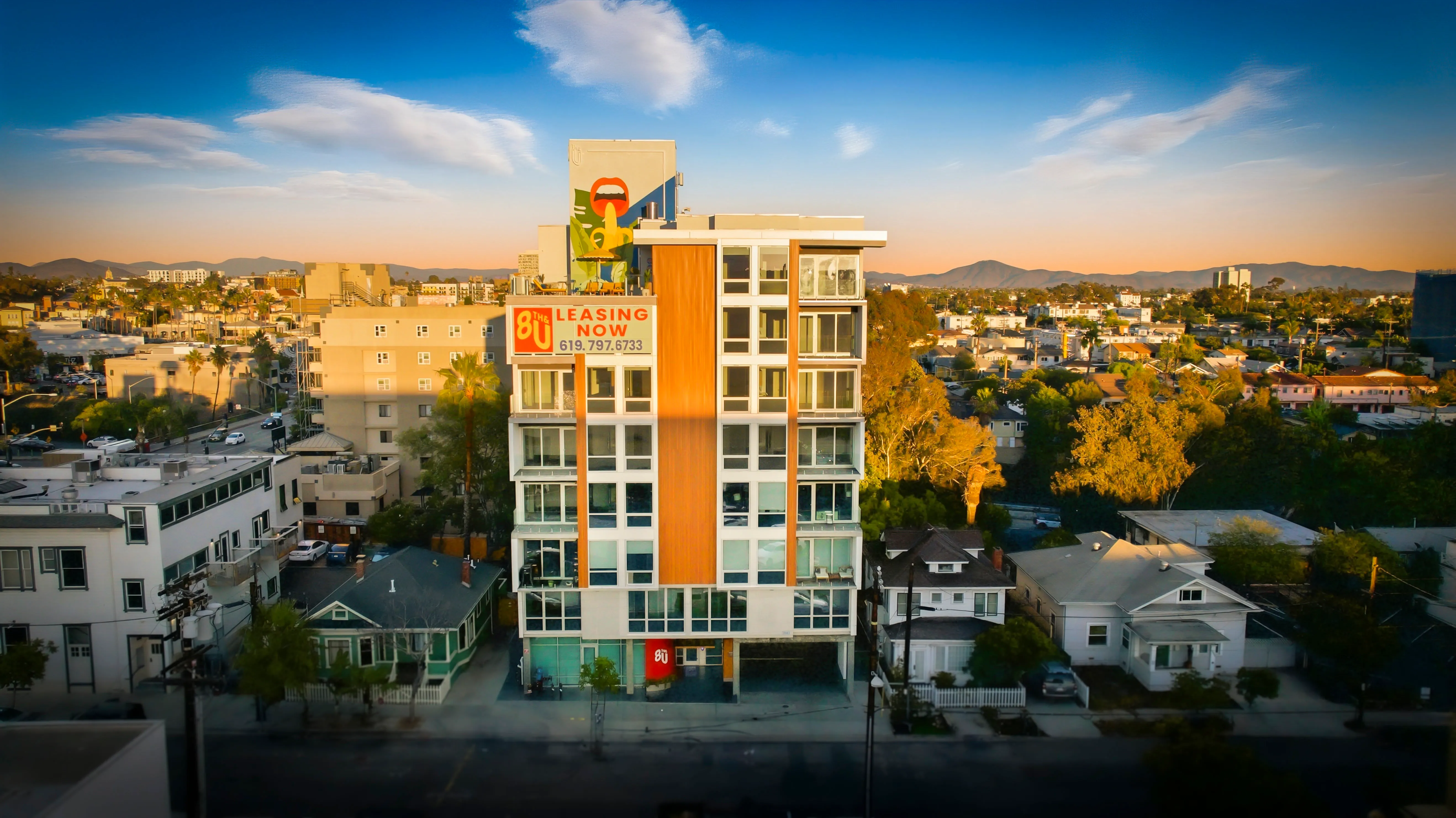 Modern multi-story apartment building with leasing sign and surrounding residential neighborhood at sunset.