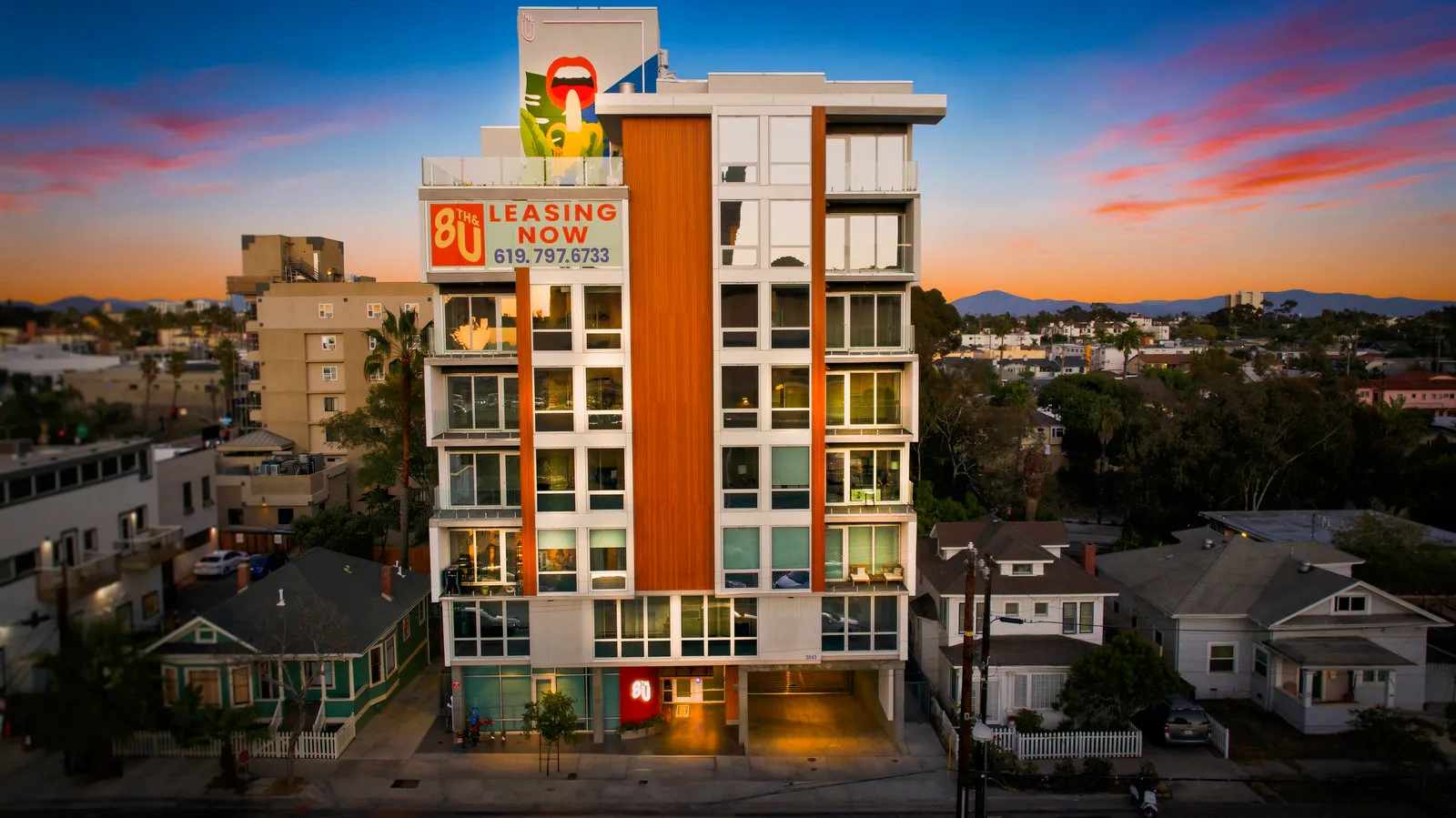 Modern apartment building with leasing sign at sunset, surrounded by smaller houses and cityscape.