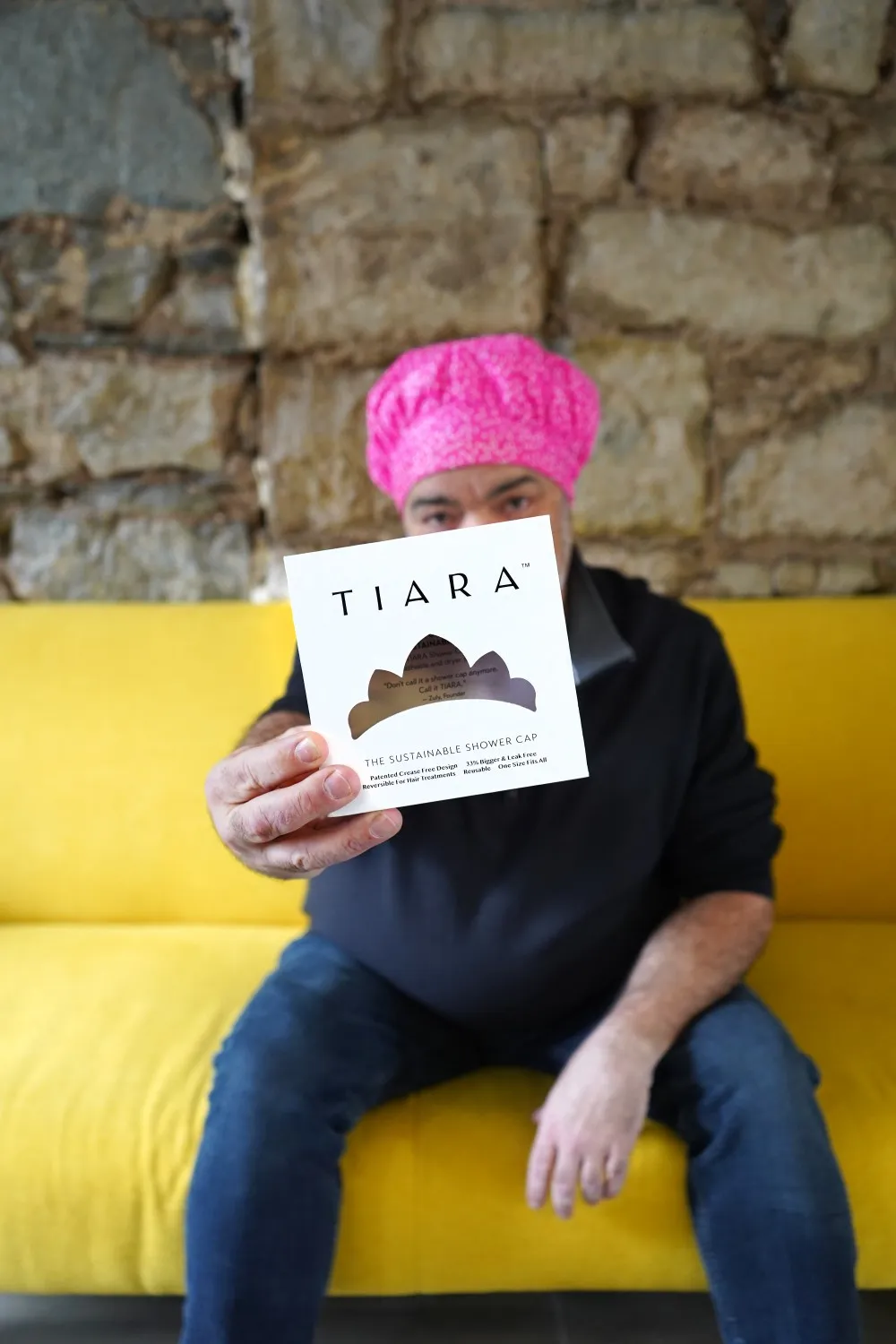 Robert Smith sitting on a yellow couch, wearing a bright pink shower cap while holding the packaging box for the shower cap (brand name: Tiara)