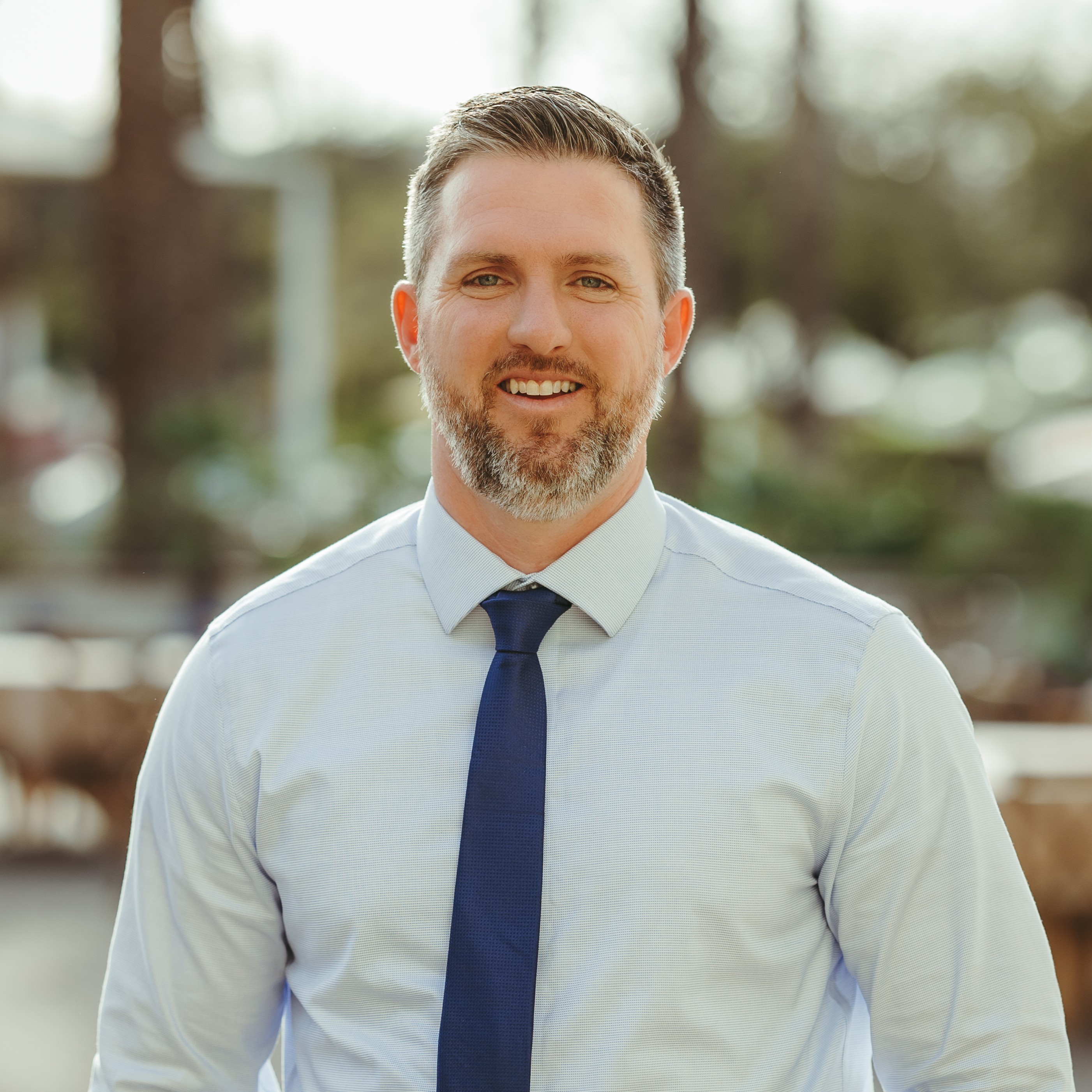 Smiling man with short hair and beard wearing a light blue dress shirt and dark blue tie outdoors.