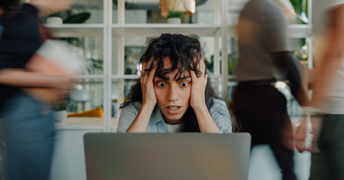A woman with curly hair looks shocked and holds her head while staring at a laptop screen, with blurred people walking by around her in an office setting.
