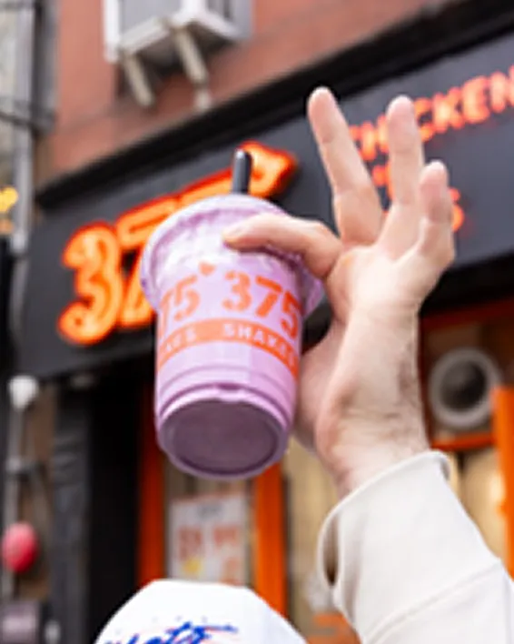 Hand holding a piece of fried chicken above a white tray with more fried chicken.