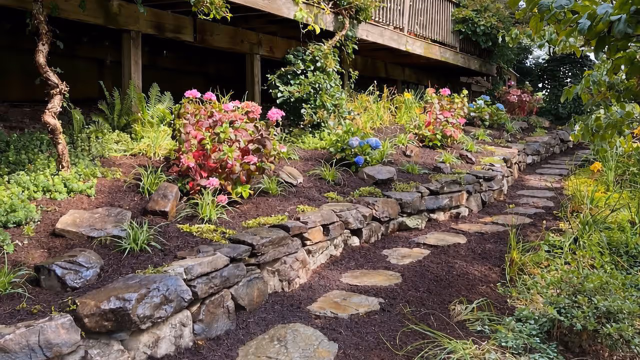 Terraced landscape design with natural stone retaining walls, decorative stepping stone pathway, and lush garden plantings beneath a raised wooden deck.