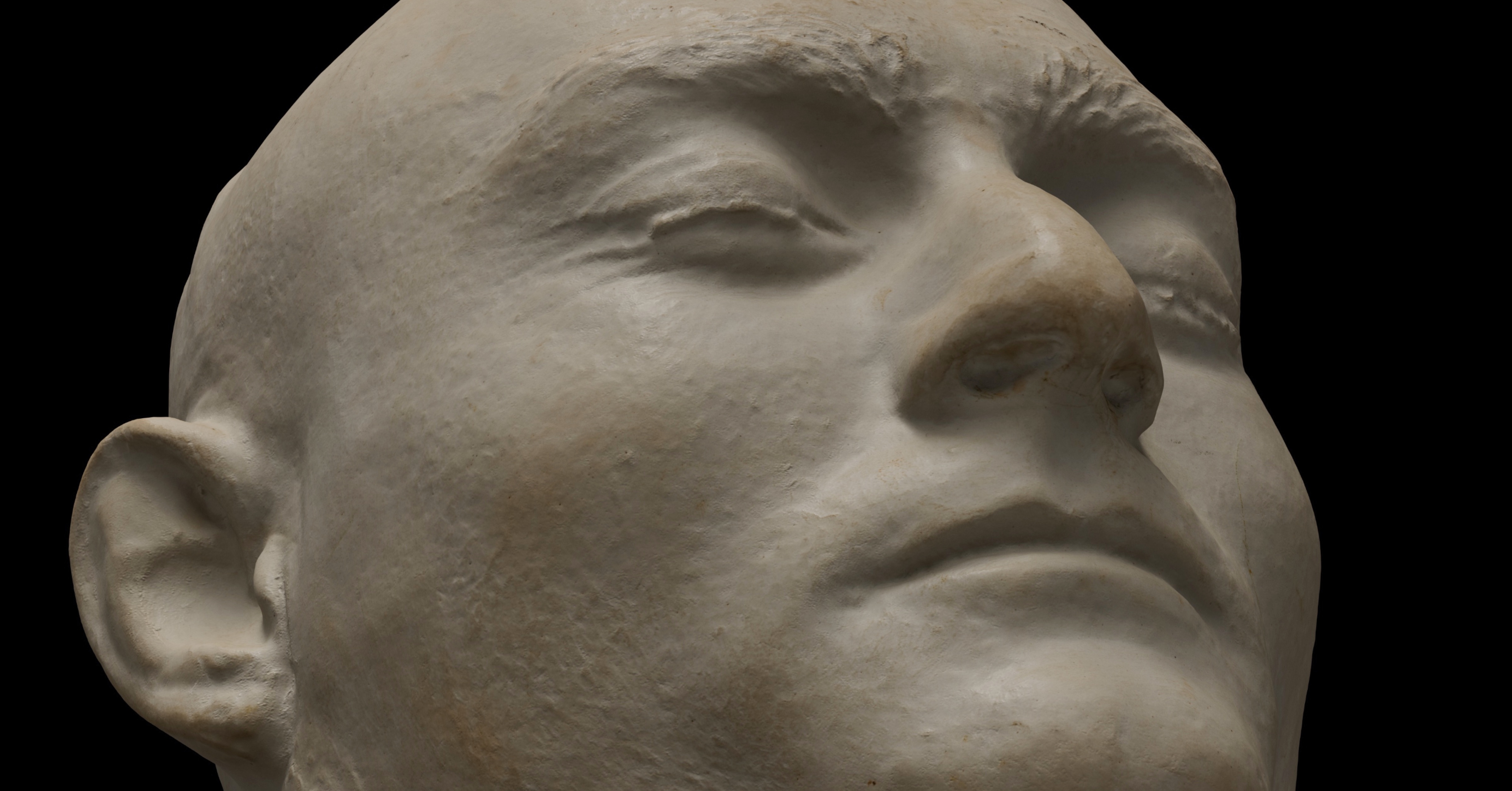 Close-up of a death mask showing a serene face with closed eyes and pronounced facial features against a black background.