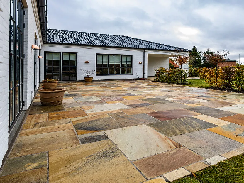 Stor terrasse belagt med champagne-farvede natursten i varierede størrelser op mod hvidt hus.