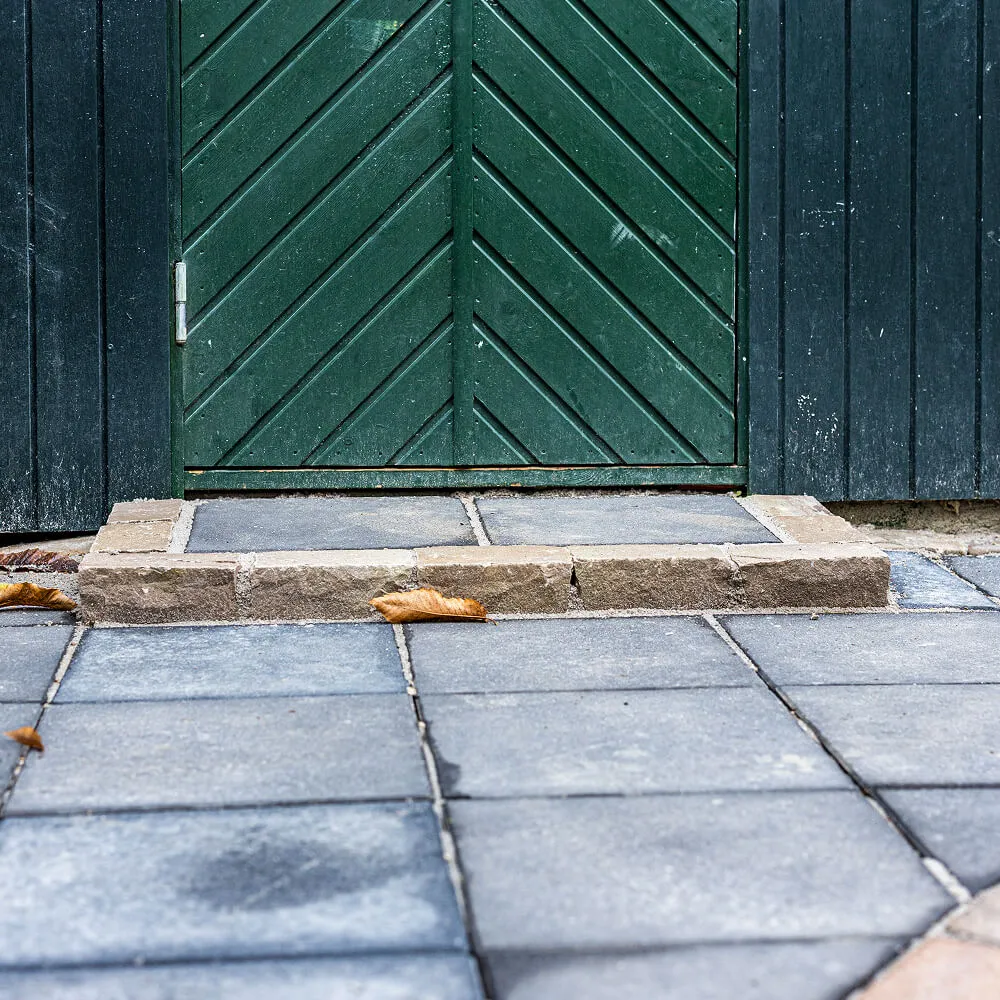 Grå flisebelagt fortov foran grøn dør med skrå paneler og små tørre blade på jorden.